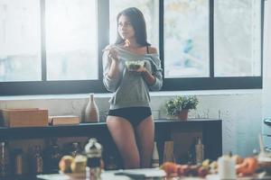 começando o dia com comida saudável. bela jovem de raça mista comendo salada e olhando para longe em pé na cozinha em casa foto