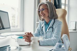 bela médica madura no jaleco branco sorrindo e desviando o olhar enquanto está sentado em seu escritório foto