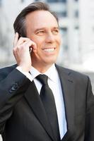 conversa de negócios. homem sênior confiante em trajes formais falando no celular e sorrindo em pé ao ar livre foto