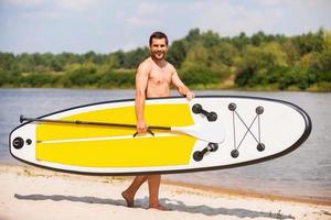 pronto para remar. jovem bonito segurando seu paddleboard enquanto caminhava pela margem do rio foto