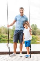 pronto para a pesca. comprimento total de pai e filho segurando varas de pescar e sorrindo em pé na margem do rio juntos foto