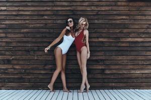 deusas do verão. comprimento total de duas mulheres jovens atraentes em trajes de banho sorrindo e se unindo enquanto posava contra a parede de madeira ao ar livre foto