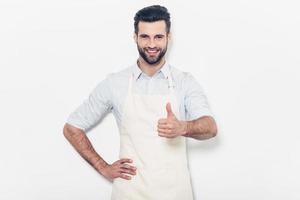 bom trabalho confiante jovem bonito no avental mantendo os braços cruzados e sorrindo em pé contra o fundo branco foto