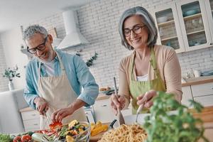 feliz casal sênior em aventais preparando um jantar saudável e sorrindo enquanto passa o tempo em casa foto