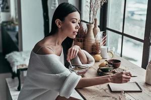 planejando o dia. mulher jovem e atraente tomando café da manhã saudável e escrevendo algo enquanto está sentado perto da janela em casa foto