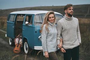 jornada feliz. lindo casal jovem de mãos dadas e sorrindo em pé perto da mini van azul estilo retrô foto