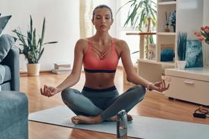 calma jovem em roupas esportivas meditando enquanto está sentado em posição de lótus em casa foto