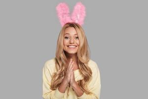sentindo animado. mulher jovem e atraente sorridente em orelhas de coelho rosa, mantendo as mãos entrelaçadas e olhando para a câmera em pé contra um fundo cinza foto