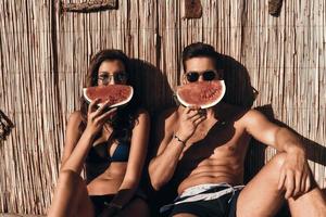 suculento e doce. lindo casal jovem cobrindo metade de seus rostos com uma fatia de melancia enquanto está sentado ao ar livre foto