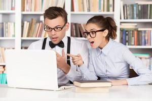 casal nerd. casal jovem nerd surpreso de óculos olhando para o monitor do computador e mantendo a boca aberta enquanto estão sentados juntos na biblioteca foto