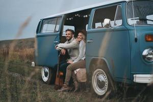 a vida é uma aventura. lindo casal jovem olhando para longe e sorrindo enquanto está sentado na mini van azul estilo retrô foto