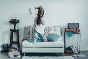 sorria mulher jovem e bonita em roupas casuais e chapéu de malha tirando uma foto por sua câmera retro em pé no sofá em casa