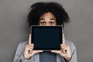 sua solução está aqui. jovem africano escondendo o rosto atrás do tablet digital com espaço de cópia em pé contra um fundo cinza foto