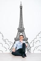 sonhando com paris. jovem bonito, mantendo a mão no queixo e olhando para longe com um sorriso enquanto está sentado no chão com ilustração da torre eiffel ao fundo foto