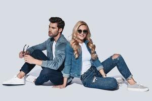 olhar na moda. lindo casal jovem vestindo jeans sorrindo enquanto está sentado contra um fundo cinza foto