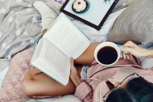 vista superior da bela jovem de pijama lendo livro e desfrutando de café da manhã enquanto descansa na cama em casa foto