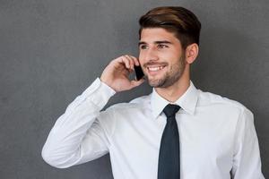 boa conversa de negócios. jovem bonito de camisa e gravata falando no celular e sorrindo em pé contra um fundo cinza foto