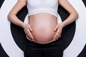 barriga de grávida. imagem recortada de mulher grávida segurando as mãos na barriga em pé contra o fundo alvo foto