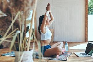 mulher jovem e bonita em roupas esportivas praticando ioga no pátio com laptop deitado perto dela foto