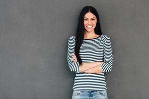 beleza confiante. mulher jovem e atraente, mantendo os braços cruzados e olhando para a câmera com sorriso em pé contra um fundo cinza foto