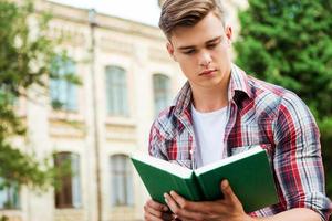 lindo leitor de livros. estudante do sexo masculino bonito lendo livro contra o prédio da universidade foto
