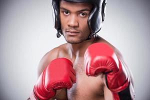 boxeador confiante. jovem africano sem camisa em luvas de boxe e capacete se preparando para lutar em pé contra um fundo cinza foto