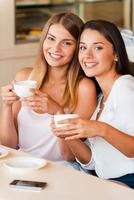 amigos no café. duas mulheres jovens atraentes segurando xícaras de café e sorrindo enquanto estão sentados no café juntos foto