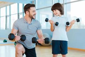 exercitando juntos. feliz pai e filho se exercitando com halteres e sorrindo em pé no health club foto