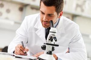 empenhado em encontrar a cura. bonito jovem cientista de uniforme branco usando microscópio e escrevendo no bloco de notas enquanto está sentado em seu local de trabalho foto