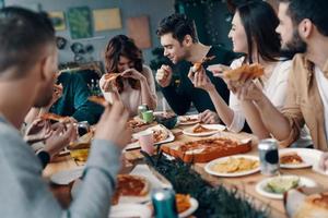 boa comida e bons amigos. grupo de jovens em roupas casuais comendo pizza e sorrindo enquanto faz um jantar dentro de casa foto