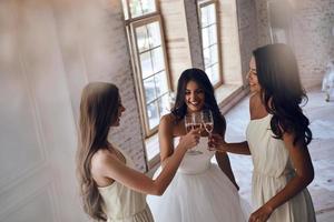 juntos em seu dia especial. vista superior da jovem noiva atraente clicando em óculos com suas lindas damas de honra em pé perto da janela juntos foto