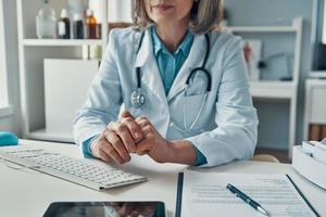 close-up da médica madura no jaleco branco, mantendo as mãos entrelaçadas enquanto está sentado em seu escritório foto