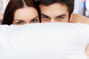 escondendo seus rostos atrás do travesseiro. alegre jovem casal deitada na cama e olhando para fora do travesseiro foto