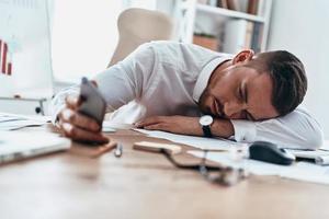 negócio é difícil. jovem cansado em trajes formais deitado na mesa e usando seu telefone inteligente enquanto trabalhava até tarde foto