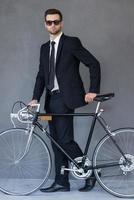 ficando verde. comprimento total do jovem empresário bonito olhando para a câmera e rolando sua bicicleta enquanto caminhava contra um fundo cinza foto