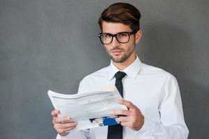 lendo algumas notícias de negócios. jovem confiante de camisa e gravata segurando jornal e olhando para a câmera em pé contra um fundo cinza foto