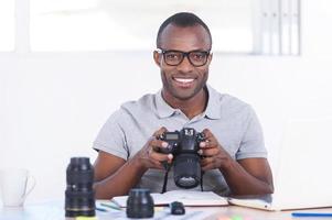 jornalista de sucesso. jovem africano bonito em roupas casuais, sentado em seu local de trabalho e segurando a câmera foto