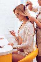 momentos de bastidores. mulher jovem e bonita usando seu smartphone com sorriso enquanto cabeleireiro escovando o cabelo na sala de maquiagem foto