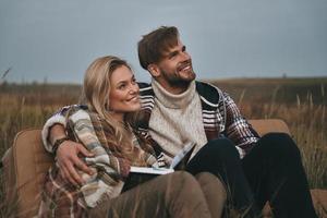 desfrutando de sua liberdade. lindo casal jovem abraçando e desviando o olhar com sorriso enquanto está sentado no campo foto