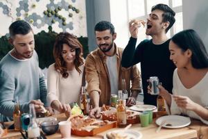 amantes de pizza. grupo de jovens em roupas casuais comendo pizza e sorrindo enquanto faz um jantar dentro de casa foto