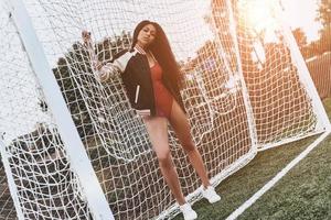 menina do futebol. comprimento total de mulher jovem e atraente em roupas esportivas, olhando para a câmera em pé perto do poste do gol foto