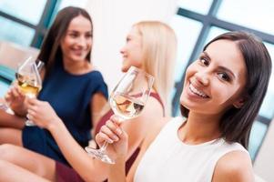 curtindo bons momentos com os amigos. bela jovem de vestido segurando o copo com vinho branco e sorrindo enquanto duas mulheres atraentes conversando ao fundo foto