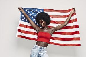 feliz jovem africana carregando a bandeira americana e sorrindo em pé contra um fundo cinza foto