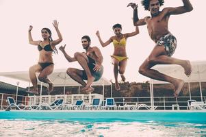 saltando para o frescor. grupo de jovens bonitos olhando felizes enquanto pulando na piscina juntos foto