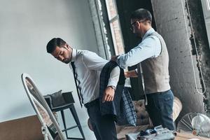 ajuste do terno. jovem designer de moda ajudando seu cliente a se vestir em pé em sua oficina foto