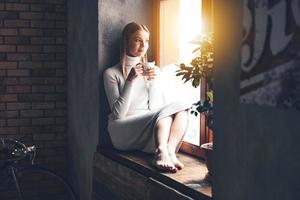 fazendo planos para um novo dia. bela jovem segurando a xícara de café e olhando pela janela enquanto está sentado no parapeito da janela em casa foto