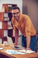 mantendo-se positivo em qualquer situação. jovem bonito olhando para a câmera e sorrindo em pé perto de seu local de trabalho no escritório foto