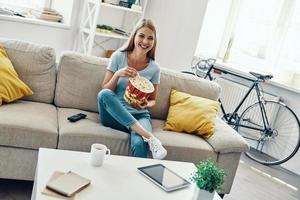 bela jovem comendo pipoca e sorrindo enquanto assiste tv no sofá em casa foto