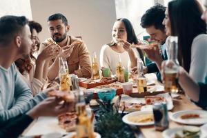 a vida é melhor com amigos. grupo de jovens em roupas casuais comendo pizza e sorrindo enquanto faz um jantar dentro de casa foto