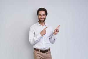 bonito jovem sorridente na camisa branca, olhando para a câmera e apontando para fora em pé contra um fundo cinza foto